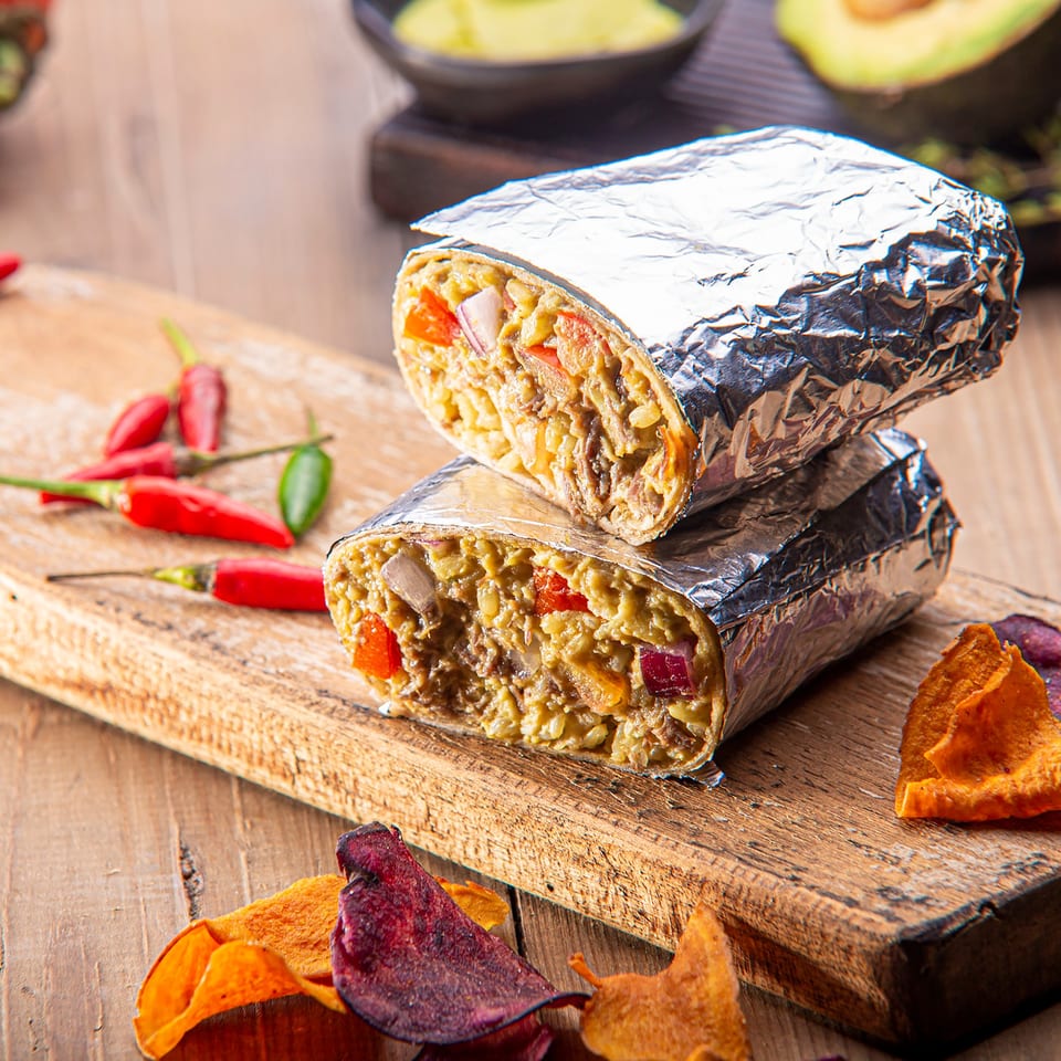 Two foil-wrapped burritos, cut in half to show fillings, on a wooden board with chili peppers.