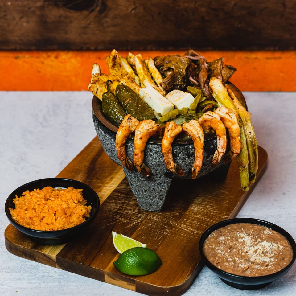 A molcajete filled with grilled shrimp, chicken, peppers, and cheese, served with rice and beans.